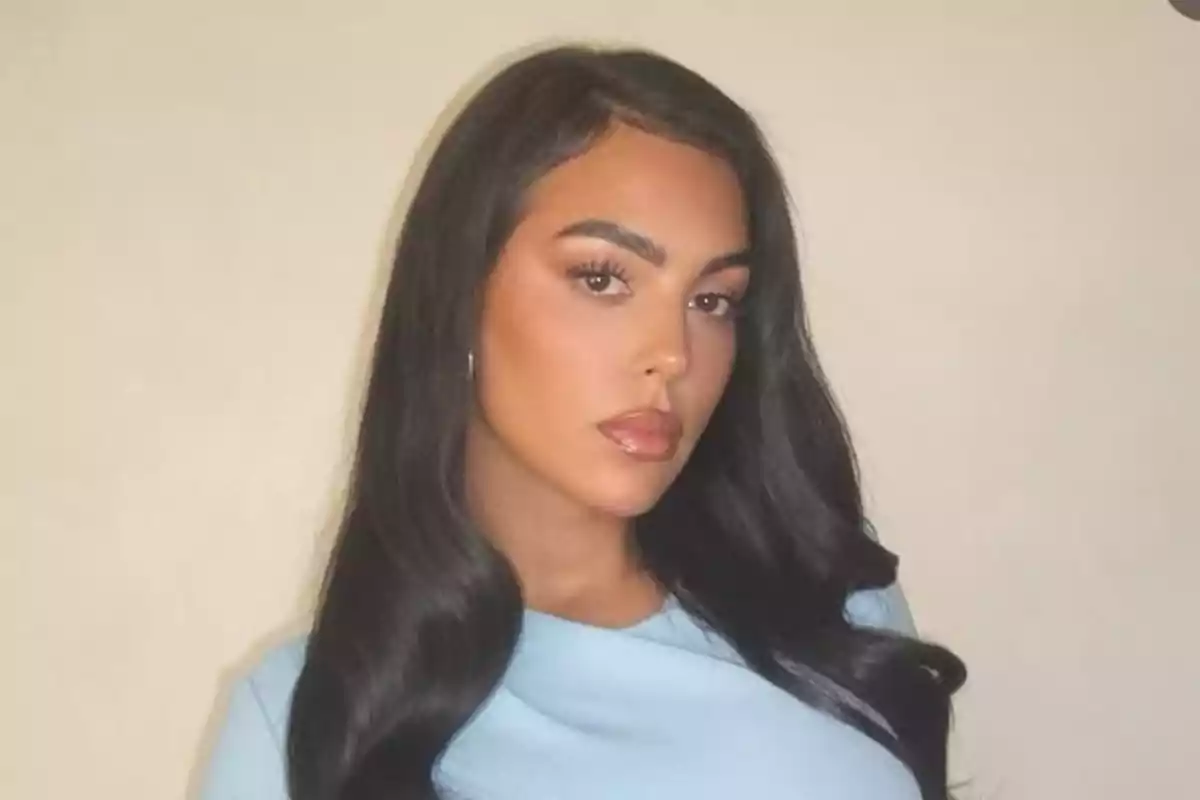 Mujer de cabello largo y oscuro con maquillaje natural y blusa azul claro posando frente a una pared beige