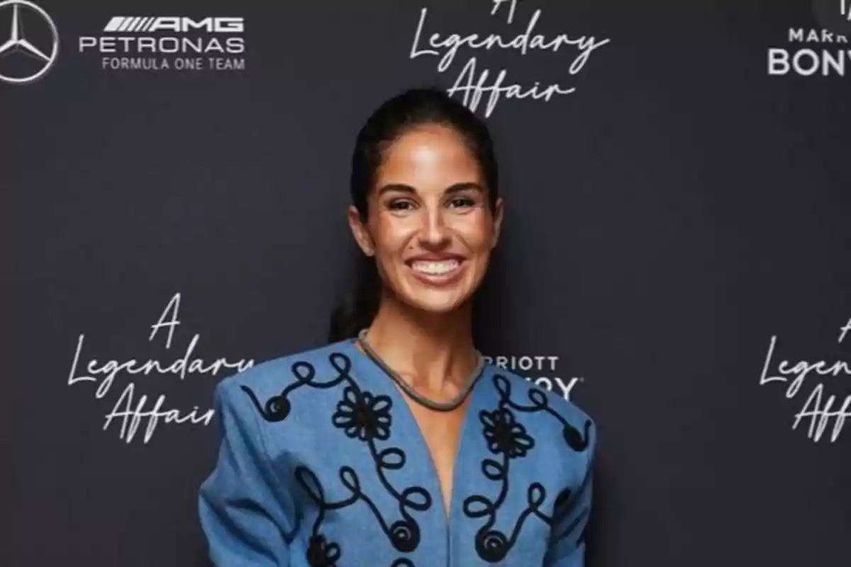 Mujer sonriendo con chaqueta azul decorada, posando frente a un fondo negro con logotipos y texto de un evento Mujer sonriendo con chaqueta azul decorada, posando frente a un fondo negro con logotipos y texto de un evento
