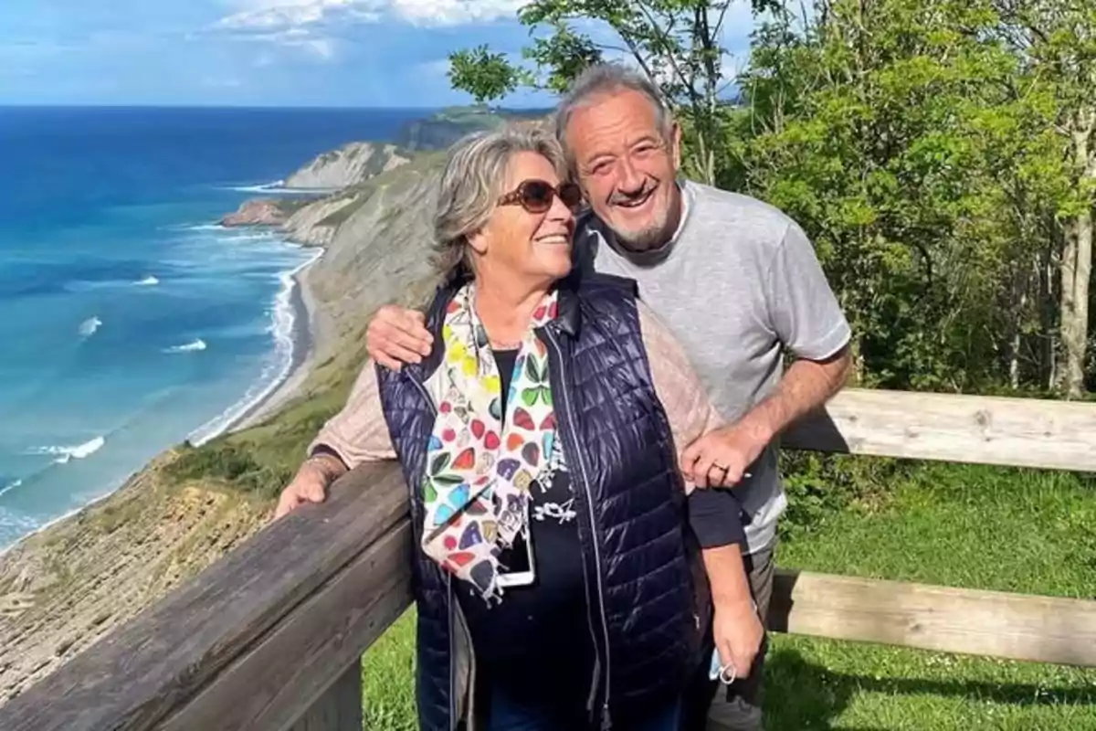 Pareja de personas mayores sonríe mientras posa junto a una barandilla de madera con un acantilado y el mar de fondo en un día soleado