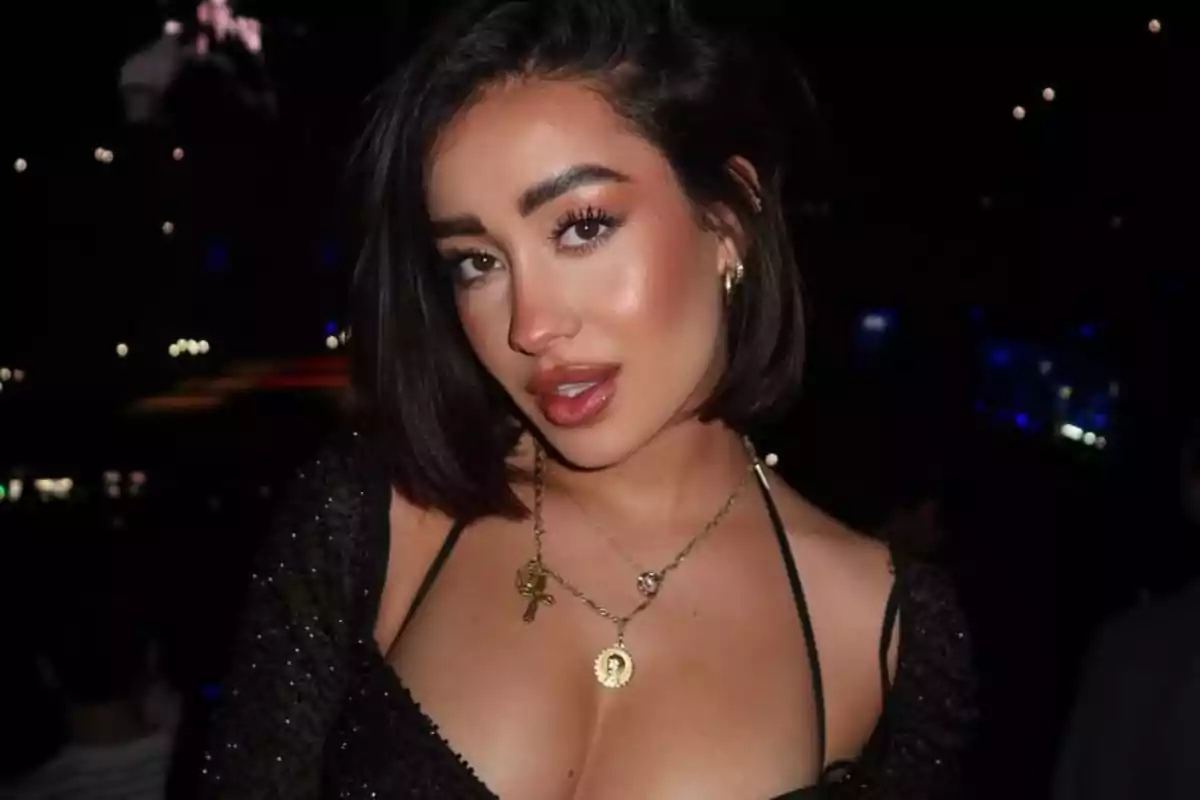 Mujer joven de cabello corto y oscuro con maquillaje llamativo, usando un vestido negro escotado y collares dorados, posando en un ambiente nocturno