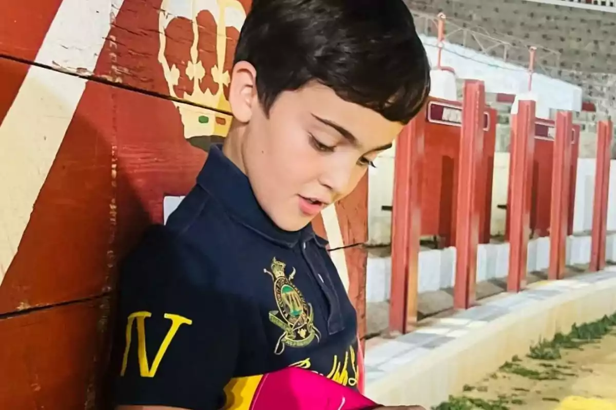 Niño de cabello corto y oscuro con camiseta azul marino apoyado en una barrera pintada de rojo y blanco en lo que parece ser una plaza de toros