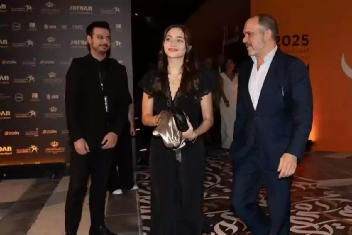 Tres personas elegantemente vestidas caminan por una alfombra en un evento con fondo de logotipos y luces cálidas