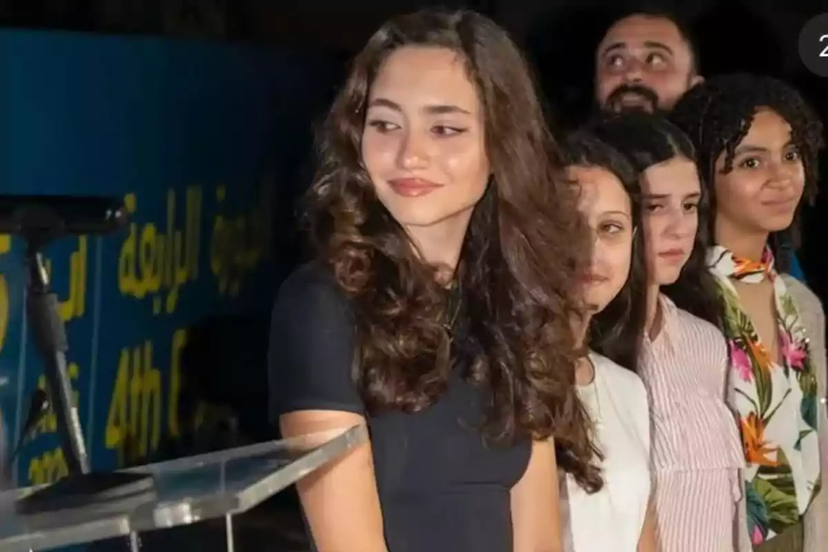 Grupo de personas jóvenes de pie en fila en un evento, con una joven de cabello rizado y vestido oscuro en primer plano mirando hacia un lado y un fondo con letras amarillas sobre azul.