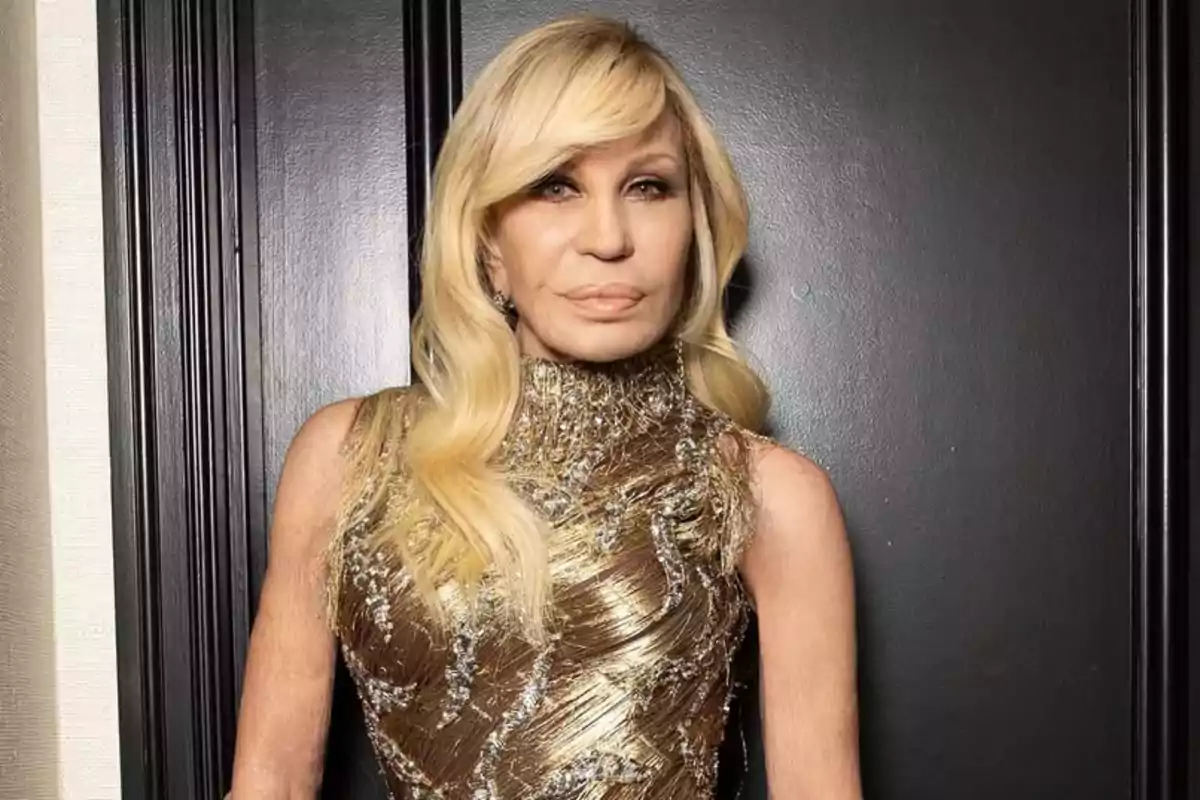 Mujer rubia con vestido dorado posando frente a una pared oscura
