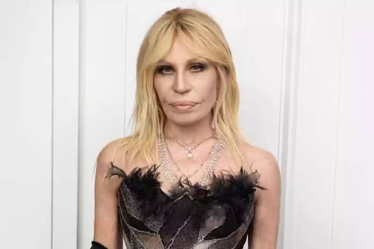 Mujer rubia con vestido negro decorado con plumas y collares plateados posando frente a una pared blanca