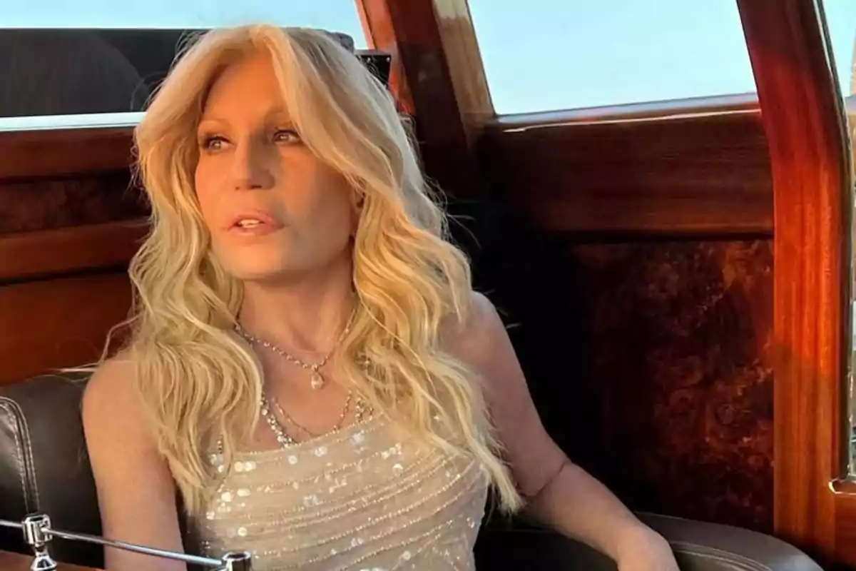 Mujer rubia con cabello largo y ondulado sentada en el interior de un vehículo de madera, usando un vestido claro y mirando hacia un lado