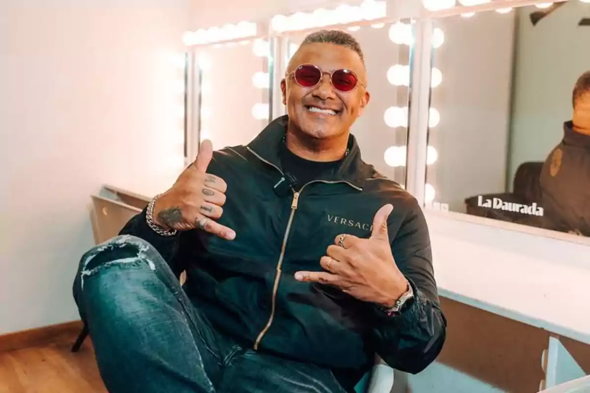 Hombre sonriente con gafas de sol rojas y chaqueta negra sentado en un camerino haciendo el gesto de shaka con ambas manos
