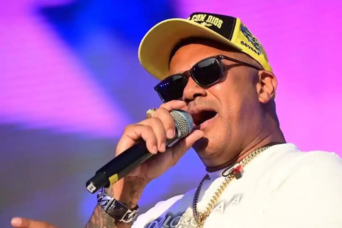 Hombre con gorra amarilla y gafas de sol cantando en un escenario con fondo colorido Hombre con gorra amarilla y gafas de sol cantando en un escenario con fondo colorido
