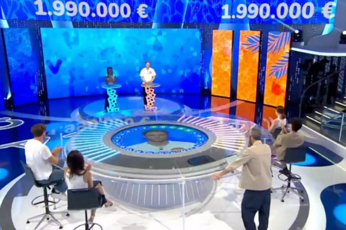 Un plató de concurso televisivo con dos concursantes de pie frente a atriles y varias personas sentadas a su alrededor mientras aplauden, con una gran pantalla azul de fondo que muestra una cantidad de dinero. Un plató de concurso televisivo con dos concursantes de pie frente a atriles y varias personas sentadas a su alrededor mientras aplauden, con una gran pantalla azul de fondo que muestra una cantidad de dinero.