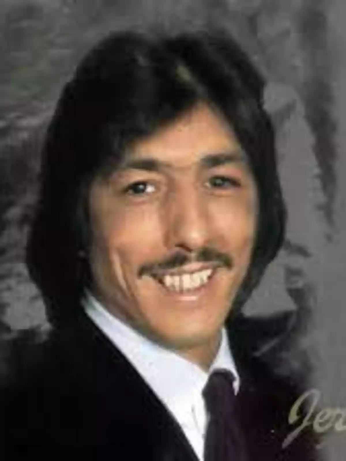 Hombre de cabello oscuro y bigote sonriendo con traje y corbata frente a un fondo gris Hombre de cabello oscuro y bigote sonriendo con traje y corbata frente a un fondo gris