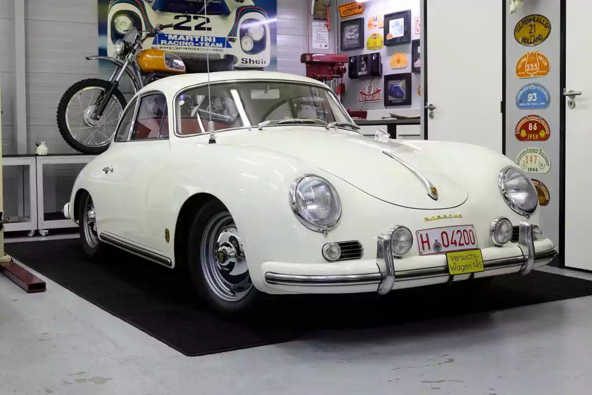 Un Porsche clásico blanco estacionado en un garaje decorado con placas y una motocicleta vintage en el fondo
