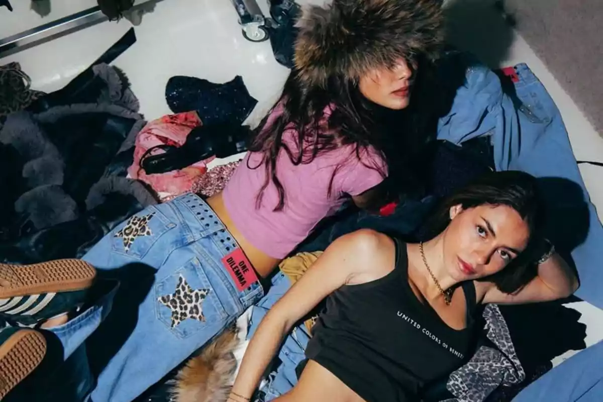 Dos chicas jóvenes posan sobre un montón de ropa en el suelo, una lleva un gorro de peluche y camiseta rosa, la otra viste un top negro y jeans claros con parches de estrellas de leopardo