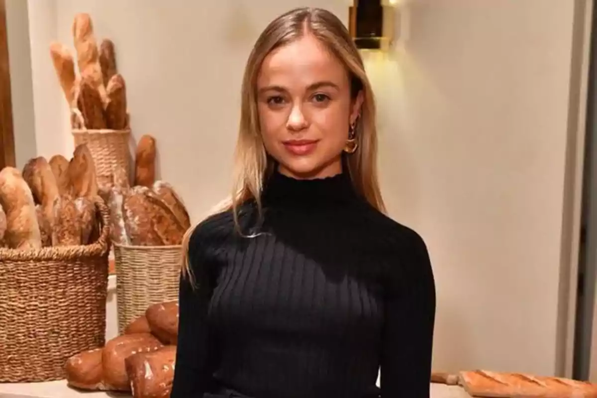 Mujer joven con cabello rubio y suelto, vestida con suéter negro, posando frente a una mesa y canastas llenas de panes en una panadería. Mujer joven con cabello rubio y suelto, vestida con suéter negro, posando frente a una mesa y canastas llenas de panes en una panadería.