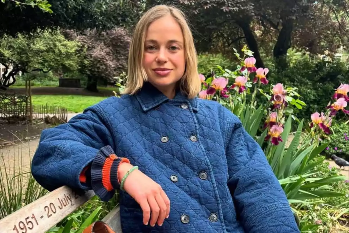 Mujer joven con cabello rubio y corto sentada en una banca de madera en un jardín con flores y vegetación al fondo lleva un abrigo azul acolchado y mira a la cámara con una expresión relajada Mujer joven con cabello rubio y corto sentada en una banca de madera en un jardín con flores y vegetación al fondo lleva un abrigo azul acolchado y mira a la cámara con una expresión relajada