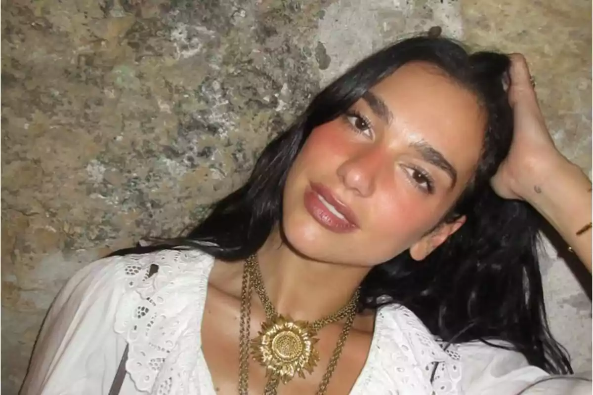 Mujer de cabello oscuro y largo con blusa blanca de encaje y collar dorado posando frente a una pared de piedra