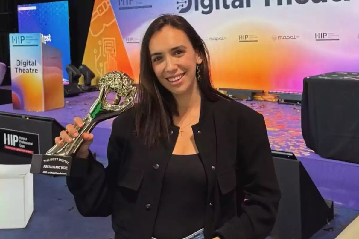 Mujer joven sonriente sostiene un trofeo en un escenario de premiación con fondo colorido y letreros de un evento profesional