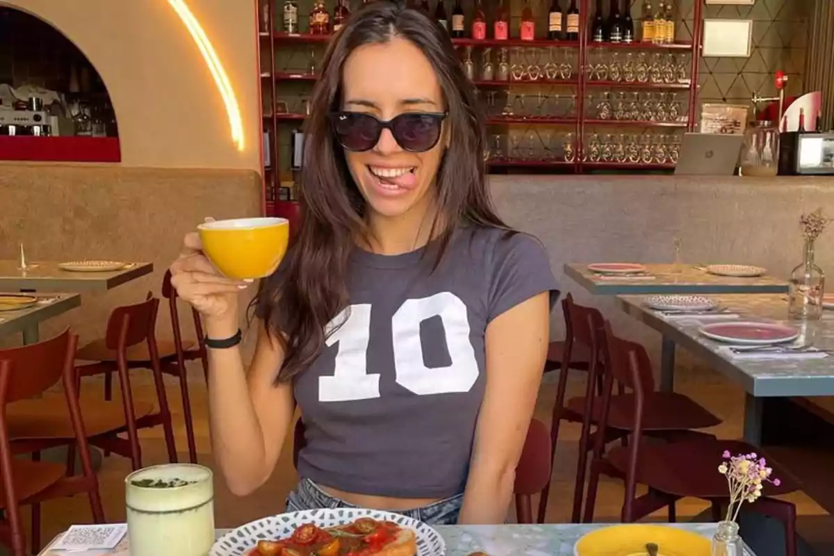 Mujer con gafas de sol y camiseta gris con el número 10 sonríe mientras sostiene una taza amarilla en una cafetería moderna con comida sobre la mesa