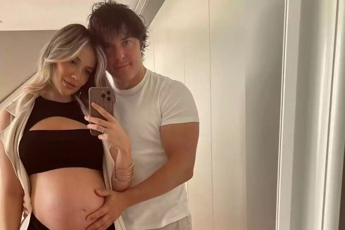 Pareja tomándose una selfie frente al espejo mientras la mujer embarazada muestra su vientre y el hombre la abraza por la cintura Pareja tomándose una selfie frente al espejo mientras la mujer embarazada muestra su vientre y el hombre la abraza por la cintura