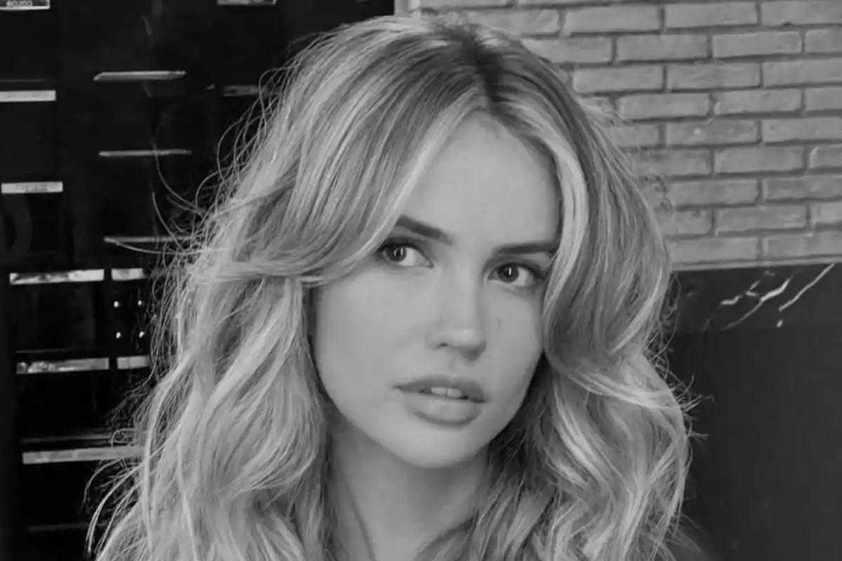 Mujer joven de cabello largo y ondulado mirando hacia un lado en una imagen en blanco y negro con fondo de ladrillos