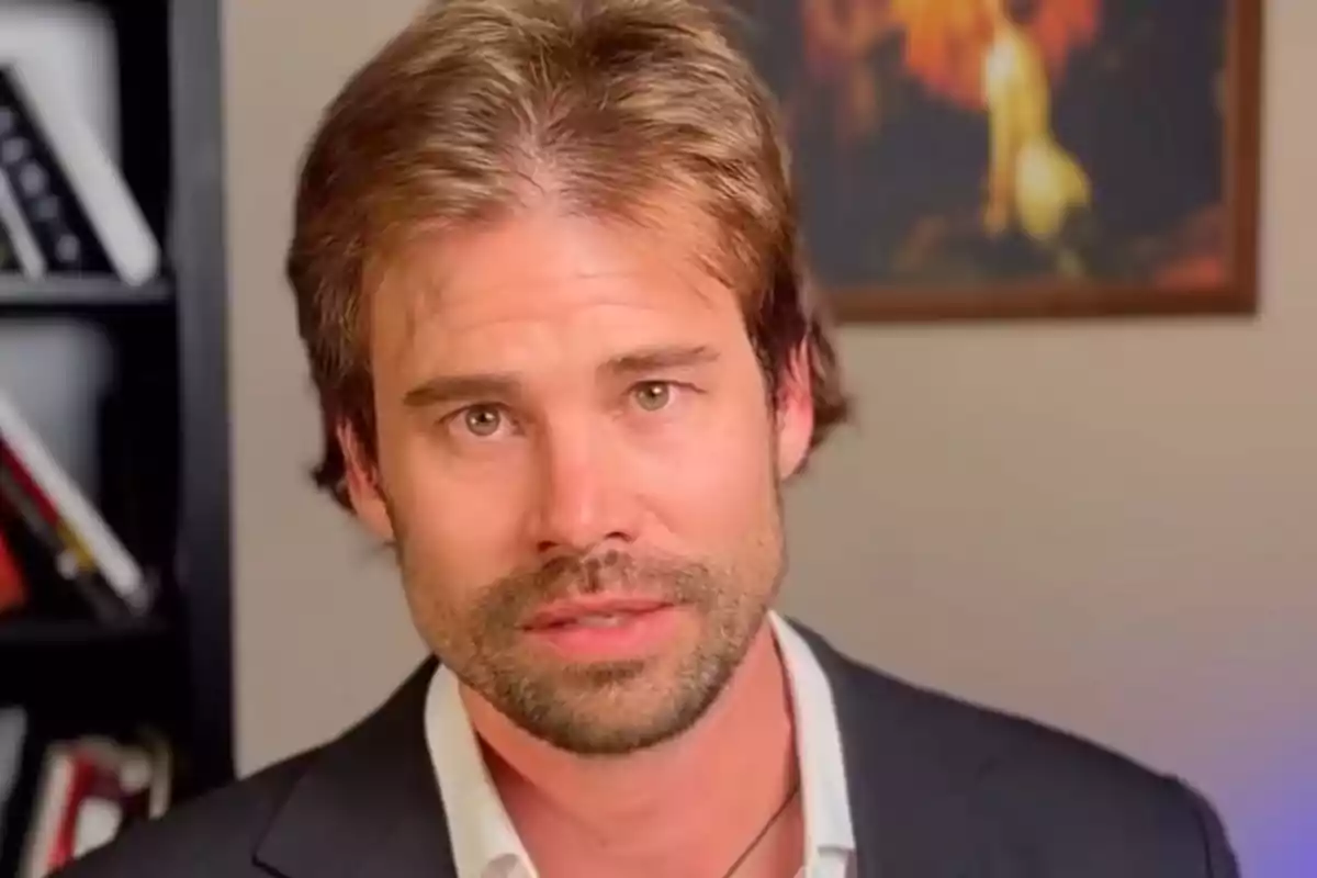 Hombre de cabello castaño claro y barba corta vistiendo saco oscuro y camisa blanca con fondo de estantería y cuadro desenfocado.