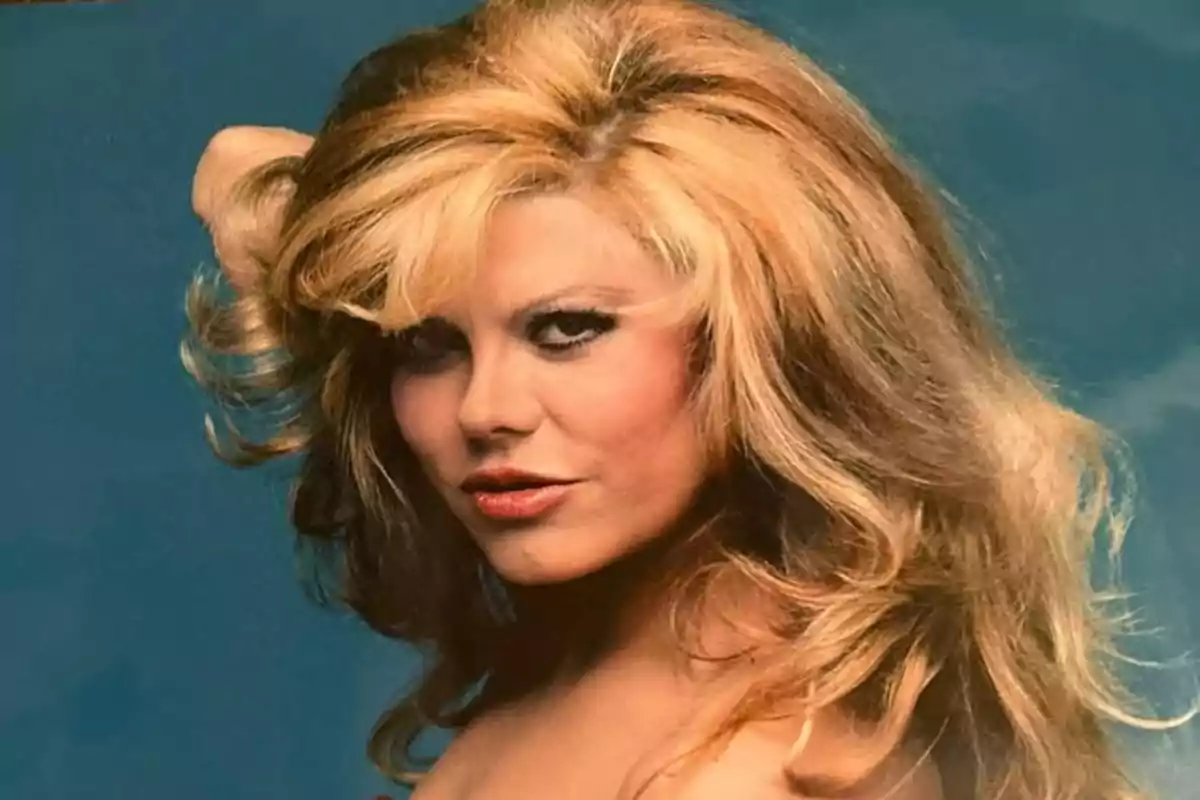 Mujer rubia con cabello voluminoso y maquillaje llamativo posando frente a un fondo azul Mujer rubia con cabello voluminoso y maquillaje llamativo posando frente a un fondo azul