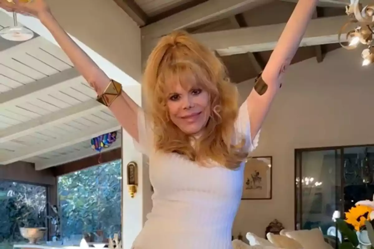 Mujer rubia con vestido blanco y brazos levantados sonríe en una sala iluminada Mujer rubia con vestido blanco y brazos levantados sonríe en una sala iluminada