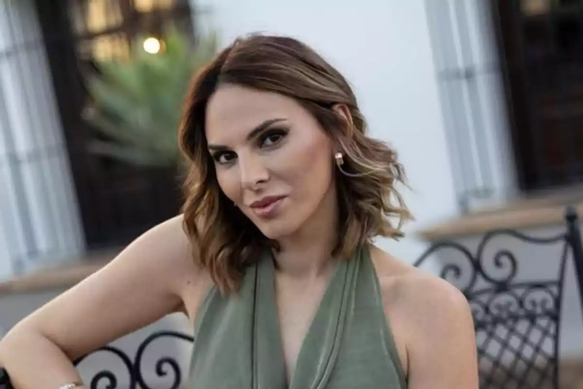 Mujer de cabello castaño claro y corto con ondas, usando un vestido verde sin mangas, sentada al aire libre frente a una estructura blanca con ventanas y rejas negras Mujer de cabello castaño claro y corto con ondas, usando un vestido verde sin mangas, sentada al aire libre frente a una estructura blanca con ventanas y rejas negras