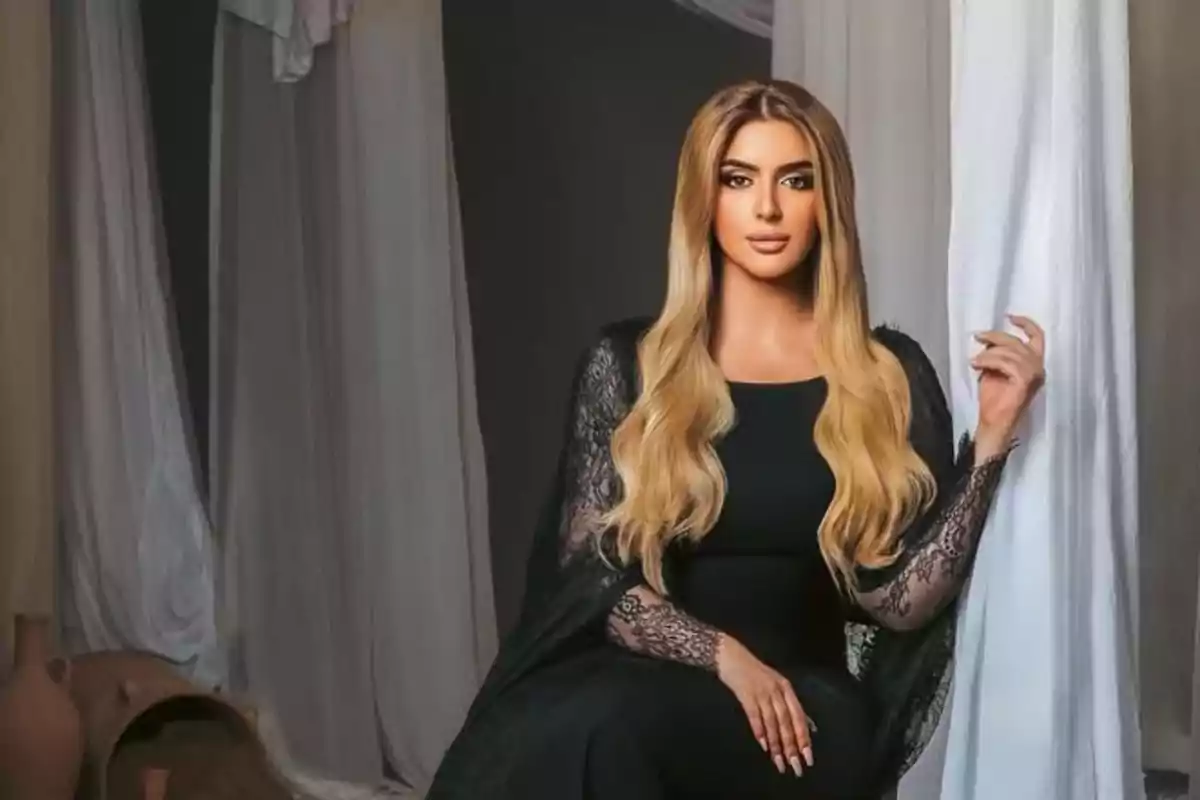 Mujer con cabello largo y rubio sentada elegantemente con un vestido negro de encaje en un ambiente decorado con cortinas blancas y jarrones de barro