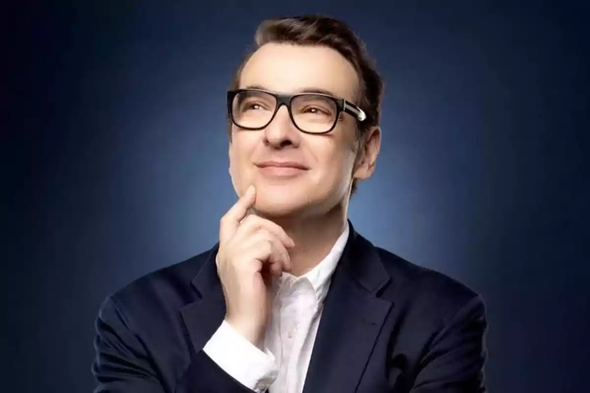 Hombre con gafas y traje oscuro sonríe mientras posa pensativo con la mano en el mentón sobre un fondo azul