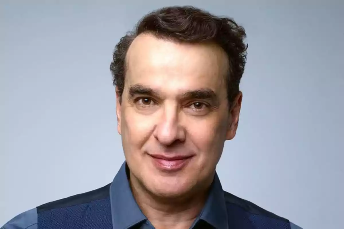 Retrato de un hombre de mediana edad con cabello corto y oscuro, vistiendo una camisa azul y sonriendo levemente frente a un fondo gris claro Retrato de un hombre de mediana edad con cabello corto y oscuro, vistiendo una camisa azul y sonriendo levemente frente a un fondo gris claro