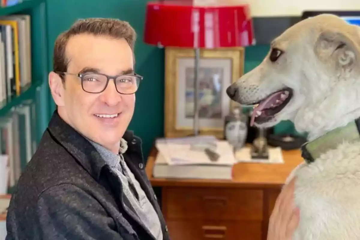 Hombre con gafas sonríe mientras acaricia a un perro grande de pelaje claro en una oficina con estantería y lámpara roja Hombre con gafas sonríe mientras acaricia a un perro grande de pelaje claro en una oficina con estantería y lámpara roja