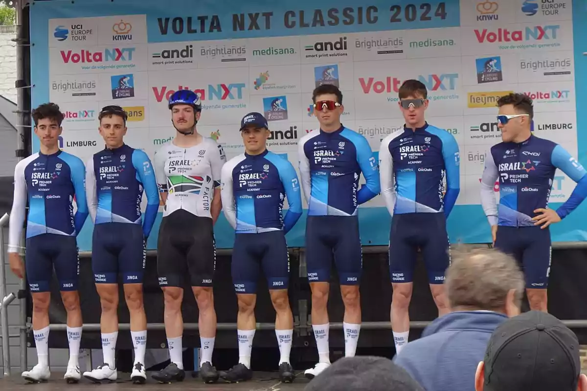 Siete ciclistas del equipo Israel Premier Tech Academy posan juntos en el podio antes de una competencia, todos con uniforme azul y blanco, frente a un fondo con logotipos de patrocinadores y el nombre del evento Volta NXT Classic 2024.