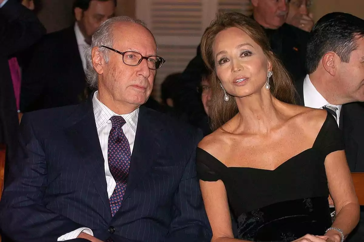 Un hombre de cabello canoso y gafas junto a una mujer de cabello largo y vestido elegante sentados en un evento formal rodeados de otras personas.