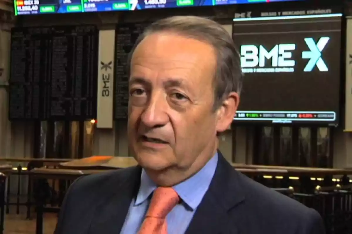 Hombre de mediana edad con traje y corbata naranja en el interior de una bolsa de valores con pantallas de información financiera al fondo