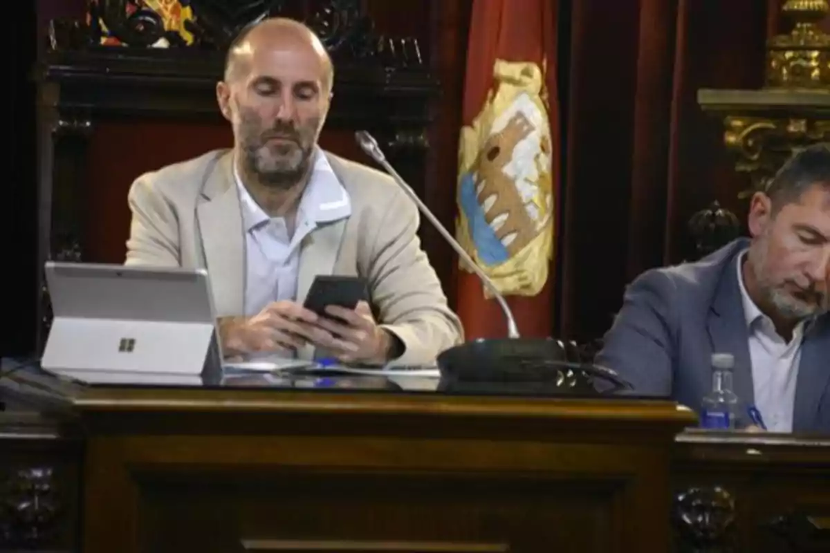 Dos hombres con saco claro y oscuro sentados en una mesa de madera, uno usando un celular y el otro escribiendo, con una bandera y una tablet en el fondo Dos hombres con saco claro y oscuro sentados en una mesa de madera, uno usando un celular y el otro escribiendo, con una bandera y una tablet en el fondo