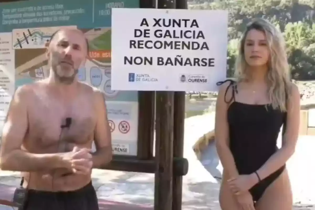 Un hombre y una mujer en traje de baño están de pie junto a un cartel que recomienda no bañarse en un área natural de Galicia Un hombre y una mujer en traje de baño están de pie junto a un cartel que recomienda no bañarse en un área natural de Galicia