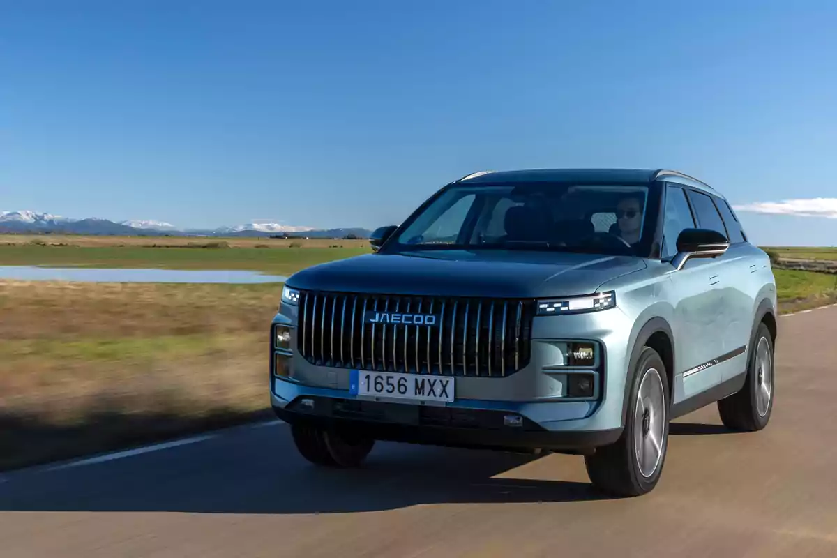 SUV Jaecoo 7 de color azul claro circulando por una carretera rural con montañas nevadas al fondo y cielo despejado SUV Jaecoo 7 de color azul claro circulando por una carretera rural con montañas nevadas al fondo y cielo despejado