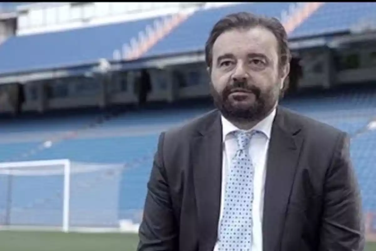 Hombre con barba y traje sentado en un estadio de fútbol con gradas azules de fondo Hombre con barba y traje sentado en un estadio de fútbol con gradas azules de fondo