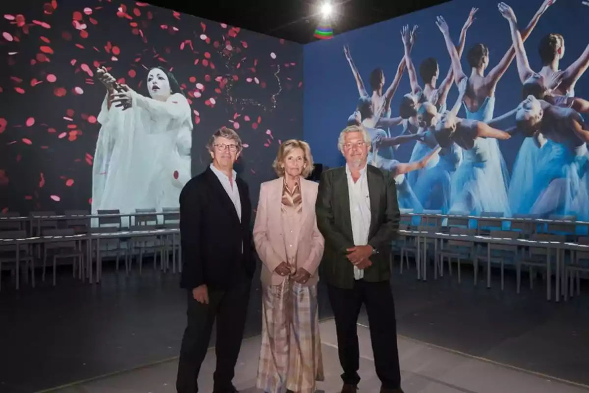 Tres personas posan en una sala con proyecciones artísticas de una bailarina y un grupo de bailarinas en el fondo Tres personas posan en una sala con proyecciones artísticas de una bailarina y un grupo de bailarinas en el fondo