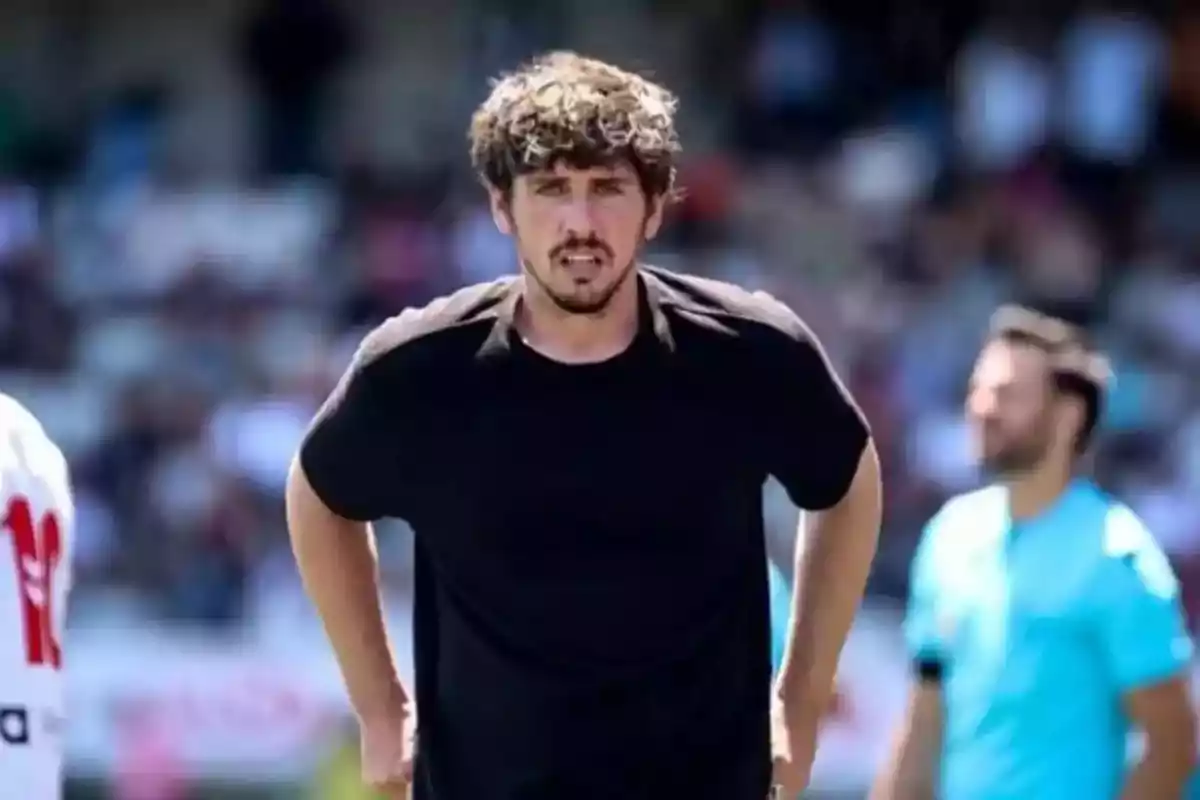 Hombre de cabello rizado y barba con camiseta negra en un campo deportivo con personas desenfocadas al fondo Hombre de cabello rizado y barba con camiseta negra en un campo deportivo con personas desenfocadas al fondo