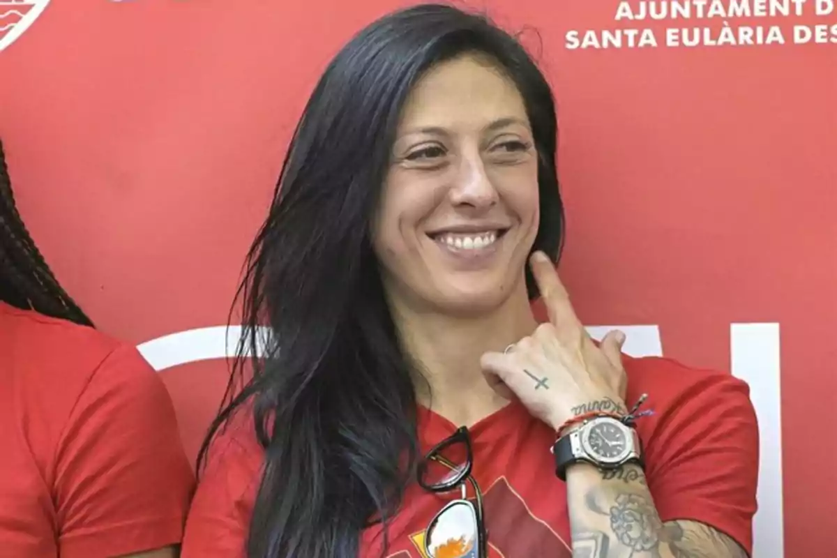 Mujer de cabello largo y oscuro sonríe mientras señala su mejilla, lleva una camiseta roja y tiene varios tatuajes en el brazo y la mano Mujer de cabello largo y oscuro sonríe mientras señala su mejilla, lleva una camiseta roja y tiene varios tatuajes en el brazo y la mano
