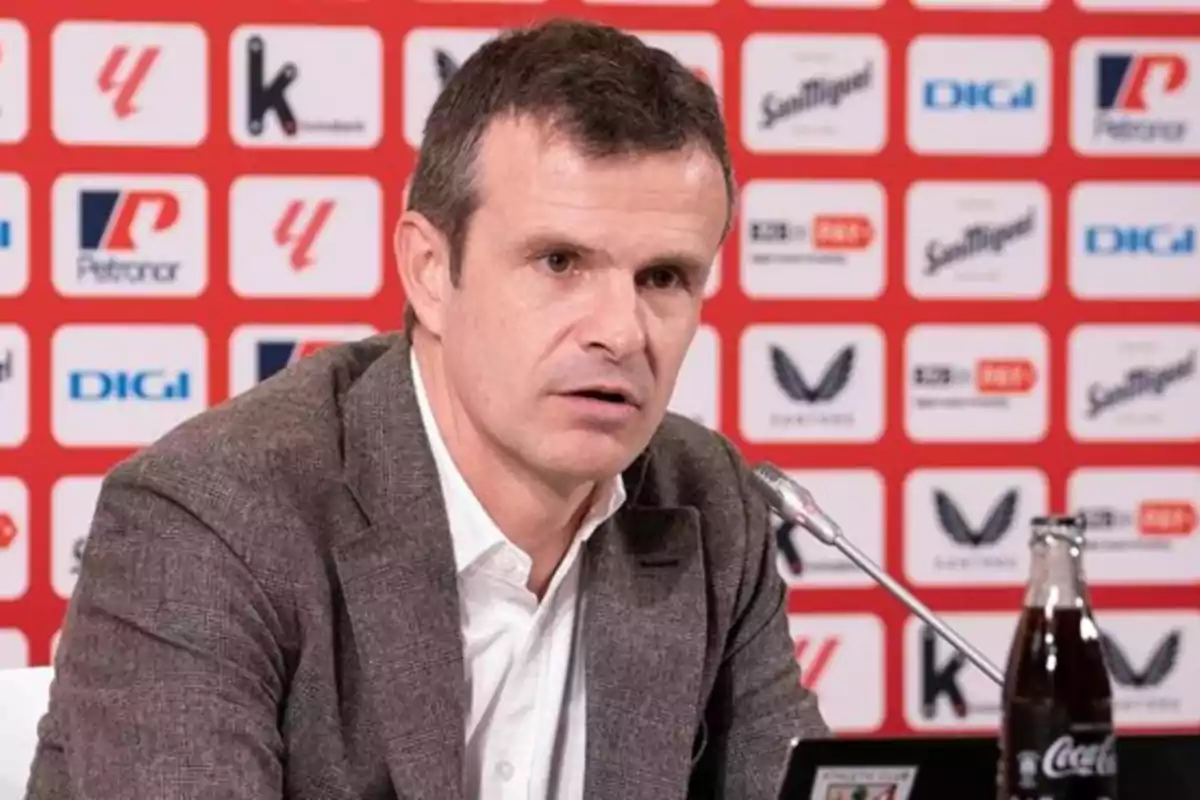 Hombre con saco marrón hablando en una conferencia de prensa con fondo de logotipos y una botella de refresco sobre la mesa Hombre con saco marrón hablando en una conferencia de prensa con fondo de logotipos y una botella de refresco sobre la mesa