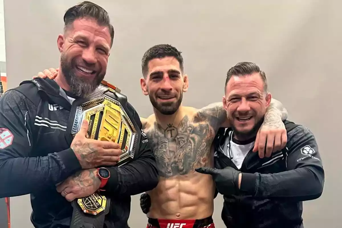 Tres hombres sonríen y posan juntos, uno de ellos sostiene un cinturón dorado de campeón de UFC y el del centro muestra el torso desnudo con tatuajes y señales de haber peleado.