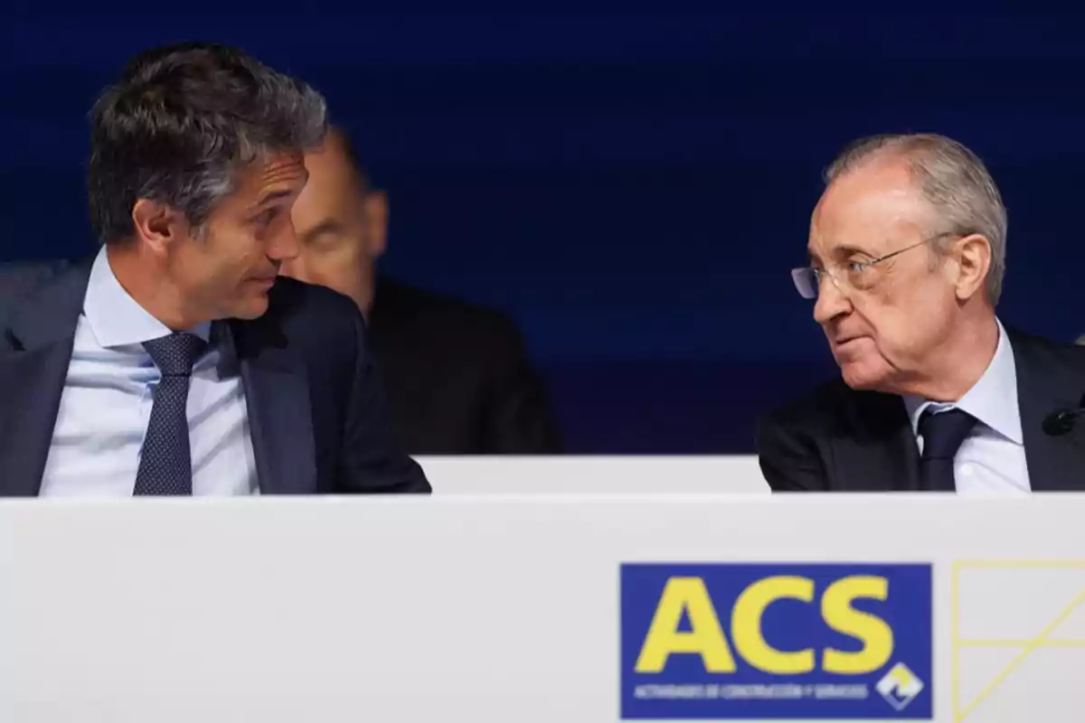 Dos hombres con traje conversan sentados detrás de una mesa blanca con el logotipo de ACS al frente Dos hombres con traje conversan sentados detrás de una mesa blanca con el logotipo de ACS al frente