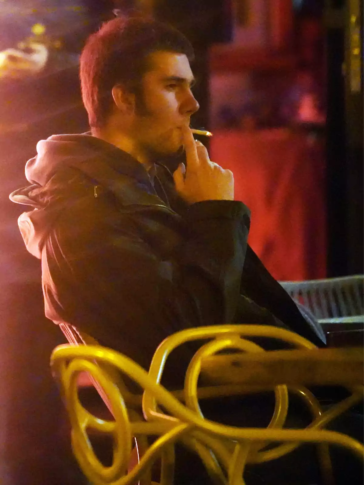 Young man sitting on a terrace at night, smoking a cigarette with a thoughtful expression Young man sitting on a terrace at night, smoking a cigarette with a thoughtful expression