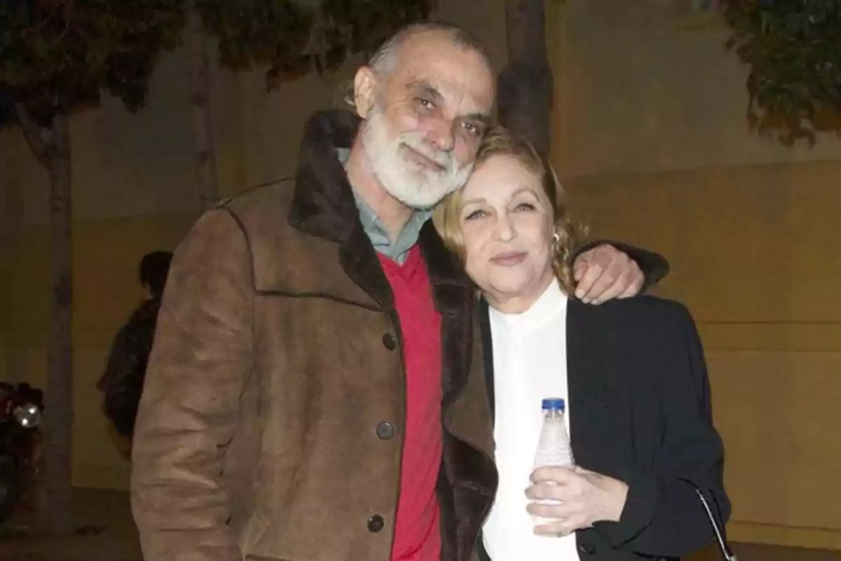 Un hombre de barba canosa abraza a una mujer rubia que sostiene una botella de agua, ambos sonríen y posan juntos al aire libre por la noche.