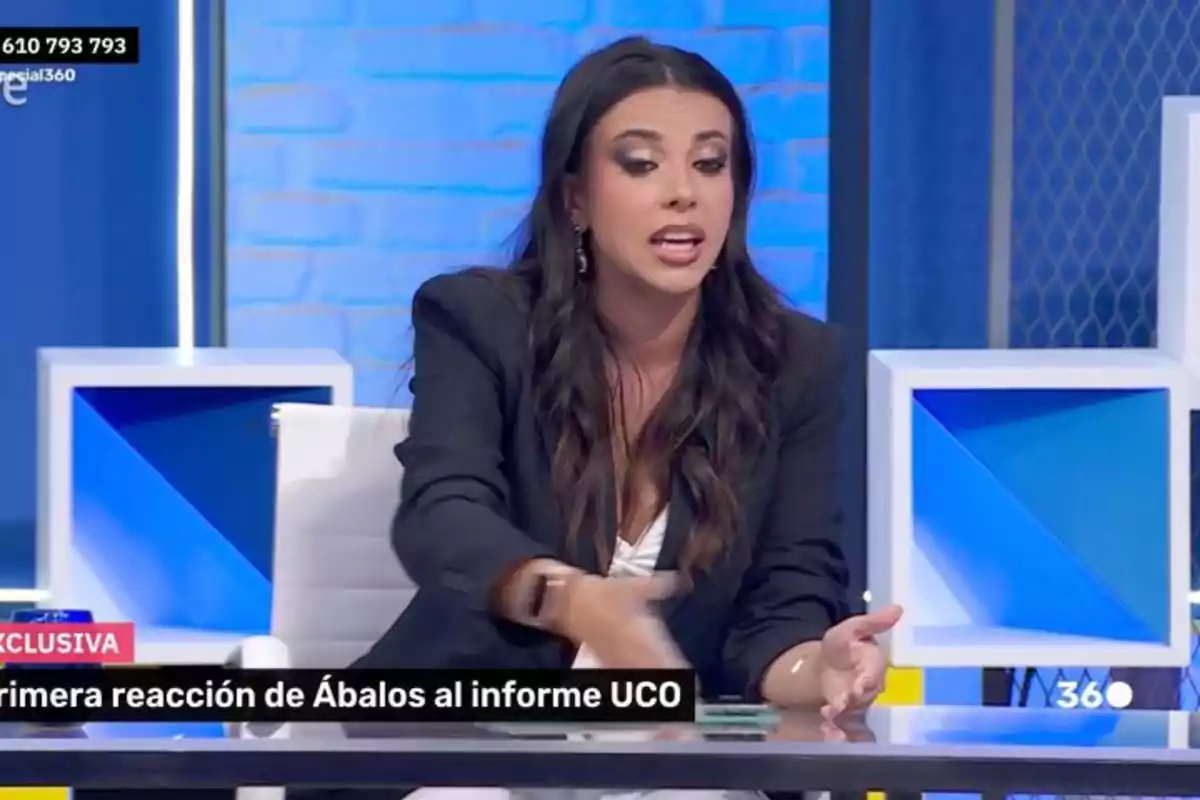 Mujer de cabello largo y oscuro sentada en un plató de televisión hablando y gesticulando con la mano sobre una mesa, con fondo azul y decoraciones geométricas. Mujer de cabello largo y oscuro sentada en un plató de televisión hablando y gesticulando con la mano sobre una mesa, con fondo azul y decoraciones geométricas.
