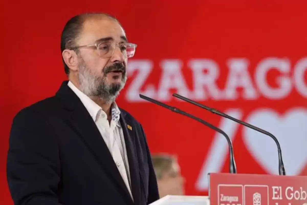 Hombre con gafas y barba hablando en un podio frente a un fondo rojo con letras blancas y un corazón Hombre con gafas y barba hablando en un podio frente a un fondo rojo con letras blancas y un corazón