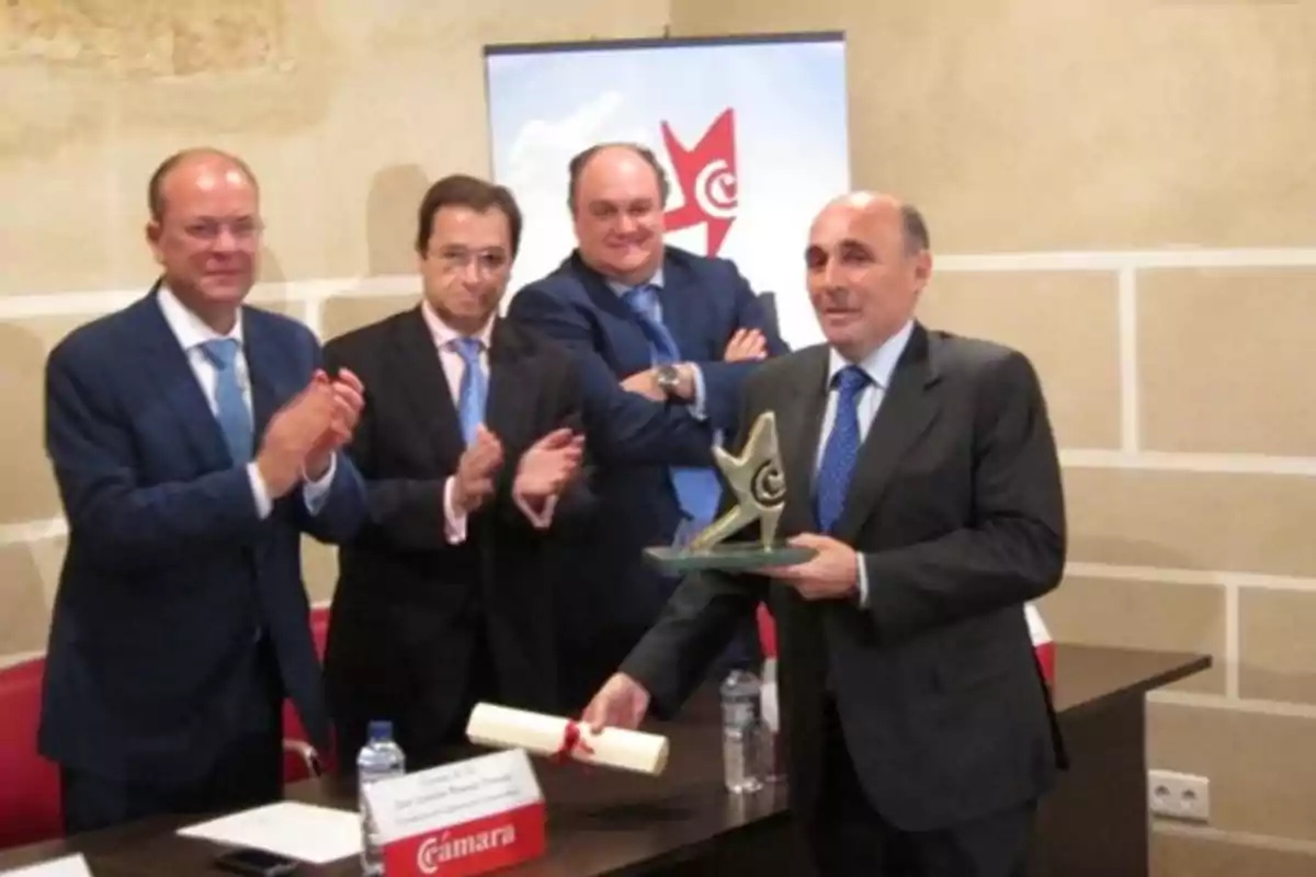 Cuatro hombres con traje en un evento formal uno de ellos sostiene un trofeo en forma de estrella y un diploma mientras los otros aplauden Cuatro hombres con traje en un evento formal uno de ellos sostiene un trofeo en forma de estrella y un diploma mientras los otros aplauden