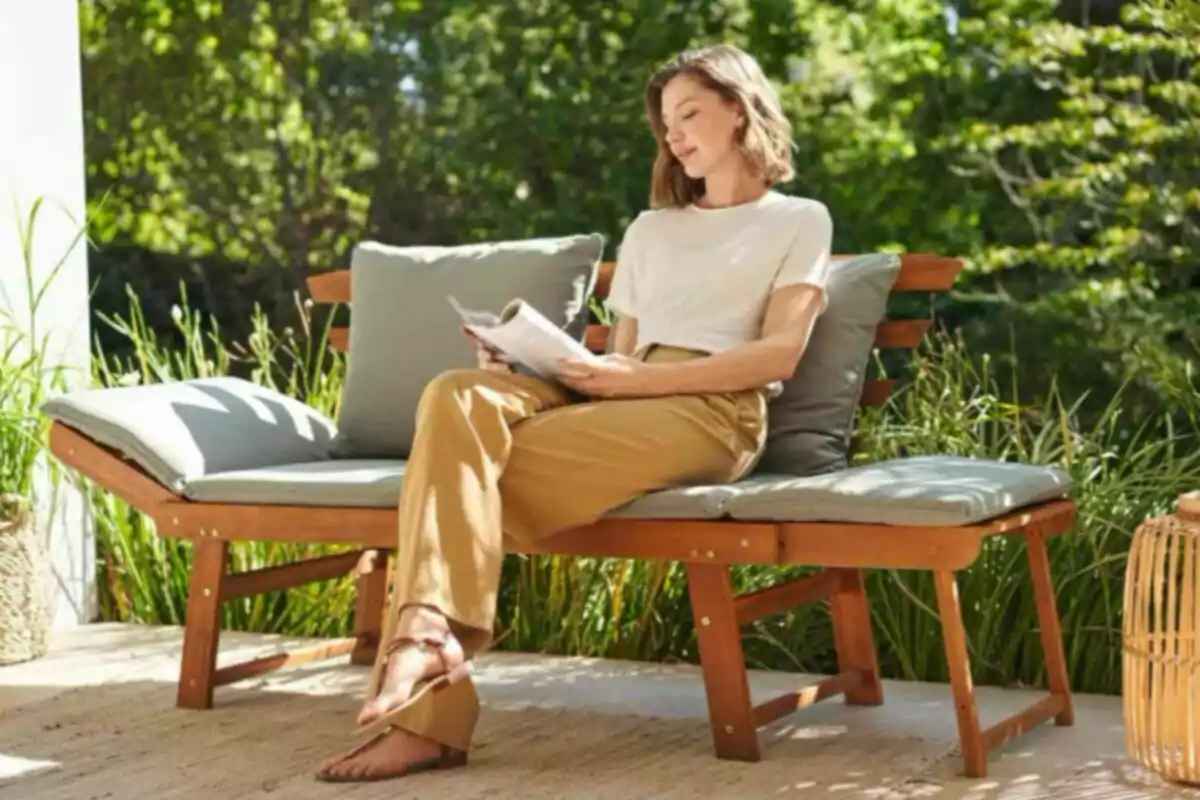 Mujer sentada en un banco de madera con cojines leyendo una revista en una terraza rodeada de plantas Mujer sentada en un banco de madera con cojines leyendo una revista en una terraza rodeada de plantas